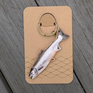 Könnte beinhalten: Ein fischförmiger Schlüsselanhänger mit einer detaillierten Illustration eines Lachses. Der Schlüsselanhänger ist an einem bronzefarbenen Ring befestigt und wird auf einer braunen Karte mit einem Wellenmuster präsentiert.