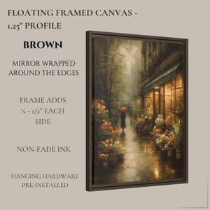May include: A floating framed canvas with a 1.25-inch profile in brown. The artwork depicts a rainy street scene with a person holding a red umbrella, surrounded by flower stalls. Features include mirror-wrapped edges and pre-installed hardware.