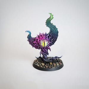 Puede incluir: Una miniatura pintada de morado y verde de una criatura con un ojo grande y un cuerpo arremolinado en forma de llama. La miniatura está sobre una base negra con textura de piedra.
