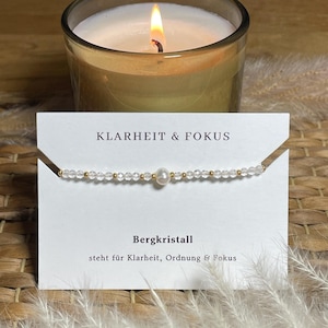 Op de afbeelding: Een delicate kralenarmband met kleine heldere gefacetteerde kristallen en kleine goudkleurige tussenstukjes, gecentreerd met een enkele witte parel. De armband wordt getoond op een witte kaart met de tekst KLARHEIT & FOKUS en Bergkristall.