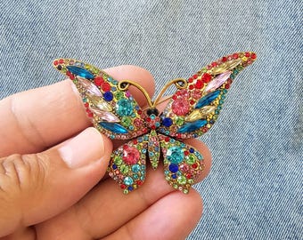 May include: A vibrant butterfly brooch with a gold-coloured metal frame. The wings are embellished with an array of sparkling gemstones in red, blue, green, pink, and yellow. The brooch is held in a hand.