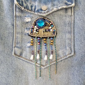 Puede incluir: Broche de medusa con cuentas con una gran gema azul en el centro, rodeada de cuentas más pequeñas en oro, azul y colores iridiscentes. El broche tiene hebras de cuentas colgantes con detalles de lágrima blanca, sobre un fondo de bolsillo de mezclilla.