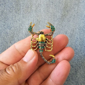 Op de afbeelding: Goudkleurige schorpioenbroche met groene en heldere kristallen accenten. Het lichaam van de schorpioen is goudkleurig, met groene kristallen op de klauwen en de staart. De broche is op een denim achtergrond.