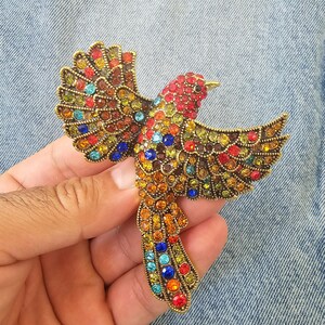 May include: A jeweled bird brooch with outstretched wings. The brooch features a red head, a body of orange and yellow, and wings with a mix of red, blue, green, and yellow stones. The bird is made of gold-toned metal.