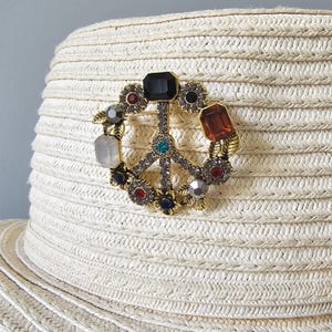 May include: A straw hat adorned with a peace sign brooch. The brooch has a gold-tone metal frame and is embellished with various coloured gemstones. The hat has a wide brim and a woven texture, typical of summer headwear.
