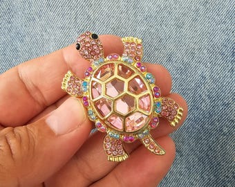 以下が含まれることがあります： ピンクと虹色のクリスタルが付いた、金色のカメのブローチ。カメの甲羅は、中央に大きなピンクのクリスタルが付いた幾何学模様です。ブローチはピンク、青、紫のアクセントがあり、手で持っています。