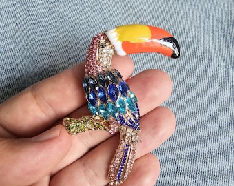 May include: A jeweled toucan brooch with a yellow, orange, and black beak. The bird's body is adorned with blue, teal, pink, and clear crystals. The toucan is perched on a gold-colored branch.