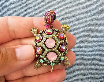 以下が含まれることがあります： 金色の金属フレームに、ピンク、赤、虹色のクリスタルをあしらった宝石付きのカメのブローチ。カメの甲羅はカラフルな宝石で飾られ、頭と足には金色のディテールが施されており、ユニークなアクセサリーとなっています。