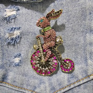 Peut inclure: Une broche en forme de lapin rose orné de bijoux, monté sur un vélo orné de bijoux. Le lapin a des yeux verts et porte un collier orné de bijoux verts. Le vélo a une grande roue ornée de bijoux et une petite roue ornée de bijoux.