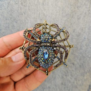 May include: A gold-toned spider brooch with a web design. The spider's body is adorned with blue and purple rhinestones, and the web features purple accents. A large, oval blue stone is centered on the spider's body.