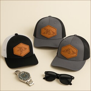 May include: Three baseball caps with leather patches and a watch and sunglasses. The caps are black and gray with mesh backs. The leather patches have a fist bump design with the words "Daddy" and "Papa". The watch has a silver band and a dark face. The sunglasses are black.