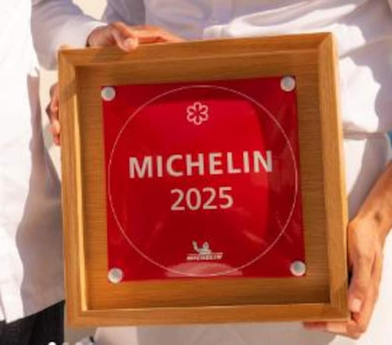 May include: A wooden framed plaque with a red centre and the text "MICHELIN 2025". The Michelin logo is at the bottom. The plaque is held by a person.