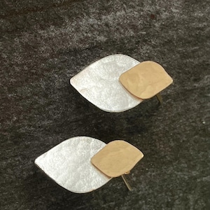 Puede incluir: Un par de pendientes plateados y dorados. Cada pendiente presenta una base plateada en forma de hoja con un acabado texturizado y un acento dorado más pequeño con textura martillada. Los pendientes se muestran sobre una superficie oscura y texturizada.