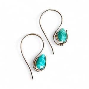 Pode incluir: Um par de brincos pendurados prateados com pedras em forma de gota de cor turquesa. As pedras são emolduradas em uma configuração enrolada em arame, e os brincos têm um design de gancho simples. O fundo é branco.