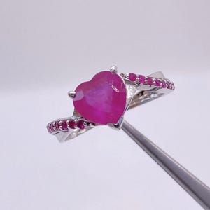 May include: A silver ring featuring a heart-shaped, vibrant pink gemstone as the centerpiece. The band is adorned with smaller, round pink gemstones. The ring is held by a silver tool against a white background.