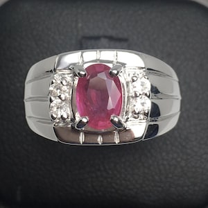 May include: A silver ring with a large, oval ruby gemstone at its centre. The ruby is a deep red hue, complemented by smaller, clear gemstones on either side. The ring has a polished, reflective finish.