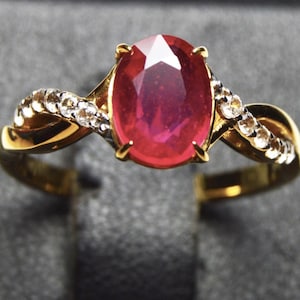 Puede incluir: Un anillo de oro con un gran rubí ovalado de color rojo intenso. El diseño del anillo es retorcido y está adornado con pequeñas piedras preciosas transparentes. El anillo se encuentra sobre un fondo gris.