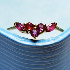 Pode incluir: Um anel de ouro com um conjunto de pedras preciosas vermelhas e rosa. O anel apresenta pedras preciosas com corte marquesa e redondas, engastadas em uma banda de ouro. As pedras são dispostas em um padrão curvo.