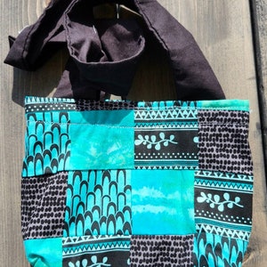 以下が含まれることがあります： 黒い布のストラップが付いた、ターコイズブルーとブラックの小さなパッチワークトートバッグ。バッグは、幾何学模様、花柄、水玉模様など、さまざまな模様の布で作られています。