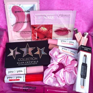 May include: A pink gift box containing beauty products. Items include an eye mask, lip mask, eyeshadow palette, lip oil, mascara, contour stick, scrunchie, and lip and eye tint. The box is lined with pink tissue paper.