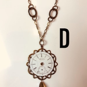 May include: A gold chain necklace with a large, ornate pendant featuring a vintage pocket watch face with Roman numerals. The watch face is cracked and has a small, clear crystal in the center. A teardrop-shaped crystal dangles from the bottom of the pendant.