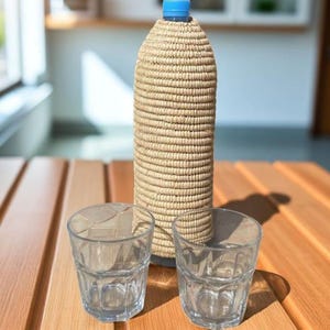 May include: A tall water bottle with a woven, beige cover and a blue cap sits on a wooden table. Two clear drinking glasses are in front of the bottle. The bottle is approximately 30 cm tall.