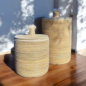May include: Two woven storage baskets with lids. The baskets are cylindrical, with a light tan color and a textured surface. The lids have decorative handles. The baskets are on a dark brown wooden surface.