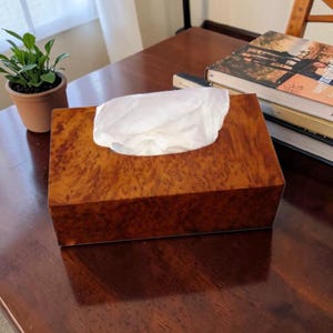 Peut inclure: Un distributeur de mouchoirs rectangulaire en bois de loupe brun, avec des mouchoirs blancs qui dépassent du dessus. La boîte est posée sur une surface en bois foncé. Une petite plante en pot et des livres sont visibles en arrière-plan.