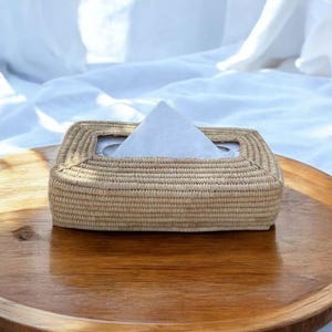 Op de afbeelding: Een rechthoekige tissuebox van geweven natuurlijke vezels, rustend op een houten dienblad. De doos heeft een vierkante opening voor het afgeven van tissues. Witte tissues zijn zichtbaar. De achtergrond is een wit laken.
