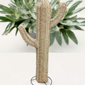 May include: A woven cactus sculpture with two arms, standing on a black metal base. The cactus is made of a light brown material with a textured, woven pattern. Green foliage is visible in the background.