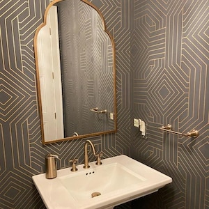 Peut inclure: Salle de bain avec un lavabo sur pied blanc, des robinets dorés et un miroir décoratif. Les murs sont recouverts d'un papier peint gris foncé avec un motif géométrique doré. Un distributeur de savon et un porte-serviettes sont également visibles.