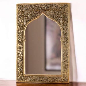 Peut inclure: Miroir rectangulaire à cadre doré, orné de motifs floraux noirs complexes. Le miroir, de forme arquée, est posé sur une table en bois, reflétant une image floue. Le design du cadre est orné et d'inspiration vintage.