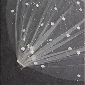 May include: White bridal veil with a single-tier design. The veil is decorated with small white flowers and pearl-like beads.