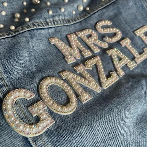 Puede incluir: Primer plano de una chaqueta vaquera azul claro con las palabras "MRS GONZALEZ" escritas con letras adornadas con perlas y pedrería. La chaqueta está decorada con perlas dispersas.