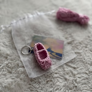 Könnte beinhalten: Ein kleiner, rosa gehäkelter Ballettschuh-Schlüsselanhänger mit Schleife. Der Schlüsselanhänger ist an einem silbernen Ring befestigt und wird auf einer weichen, strukturierten Oberfläche präsentiert. Ein kleiner Knäuel rosa Garn ist ebenfalls sichtbar.