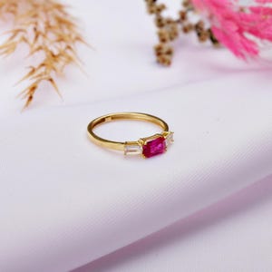 Può includere: Un anello d'oro con una pietra quadrata rosa e due pietre rettangolari trasparenti più piccole.
