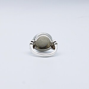 May include: A silver ring with a large, oval, gray stone. The ring has a simple band and decorative elements on either side of the stone. The stone appears to be smooth and polished, set in a silver bezel.