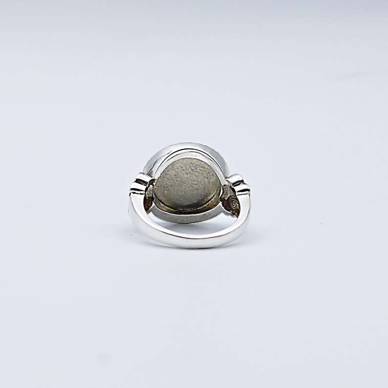 May include: A silver ring with a large, oval, gray stone. The ring has a simple band and decorative elements on either side of the stone. The stone appears to be smooth and polished, set in a silver bezel.