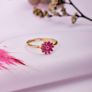 May include: A gold ring with a pink flower-shaped gemstone in the center. The ring is on a white surface.