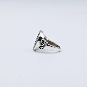 May include: A silver ring with an oval, dark-colored stone. The ring has a decorative design on the sides and a textured edge around the stone. The ring is set against a white background.