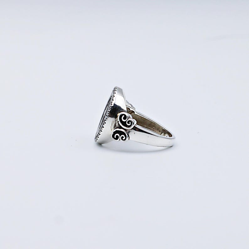 May include: A silver ring with an oval, dark-colored stone. The ring has a decorative design on the sides and a textured edge around the stone. The ring is set against a white background.
