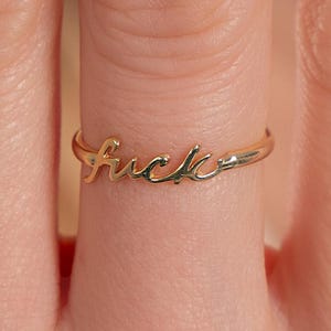 Könnte beinhalten: Goldring mit dem Wort "fuck" in Kursivschrift.