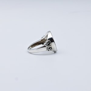 May include: A silver ring with an oval, dark-colored stone. The ring has ornate scrollwork on the band and around the stone setting. The ring is shown on a white background.