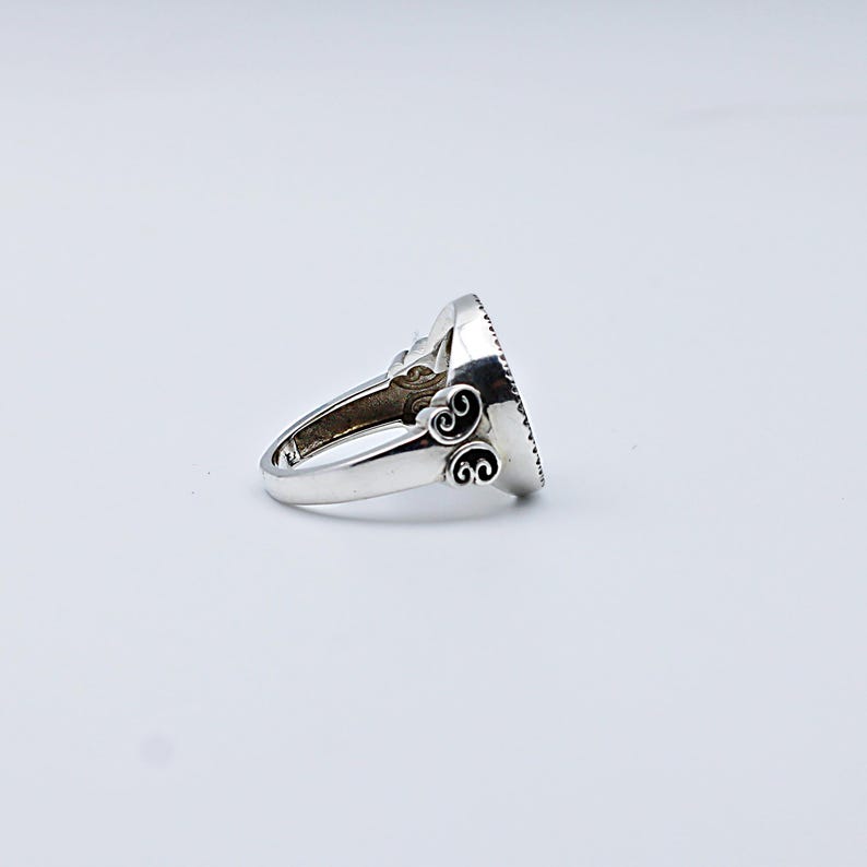 May include: A silver ring with an oval, dark-colored stone. The ring has ornate scrollwork on the band and around the stone setting. The ring is shown on a white background.