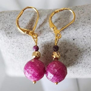 May include: A pair of gold leverback earrings. Each earring has a faceted, round, magenta bead, a gold textured bead, and a small, dark purple bead. The earrings are set against a light grey background.