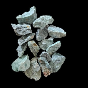 Puede incluir: Una colección de diez piedras verdes y grises, algunas con una textura rugosa, dispuestas en una pila sobre un fondo negro.