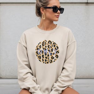 Può includere: Felpa beige con un motivo leopardato a forma circolare. La parola "HAPPY" è stampata in blu all'interno del motivo leopardato. La felpa ha maniche lunghe e girocollo.