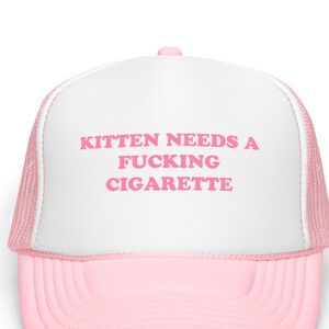 May include: A pink and white trucker hat with the text "KITTEN NEEDS A FUCKING CIGARETTE" in pink block letters. The hat has a pink brim and a white front panel with a pink mesh back.