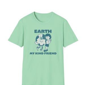 May include: A light green short-sleeve t-shirt featuring a dark blue graphic of a smiling planet Earth standing next to a person. The text above the graphic reads EARTH and the text below the graphic reads MY KIND FRIEND in bold lettering.
