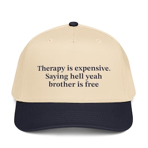 May include: A baseball cap with a cream-colored crown and a navy blue brim. The cap features the text "Therapy is expensive. Saying hell yeah brother is free" in dark blue lettering.